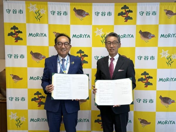 守谷市のロールアップバナーを背景に、守谷市長と神谷代表取締役が協定書を手に持ち記念撮影している様子